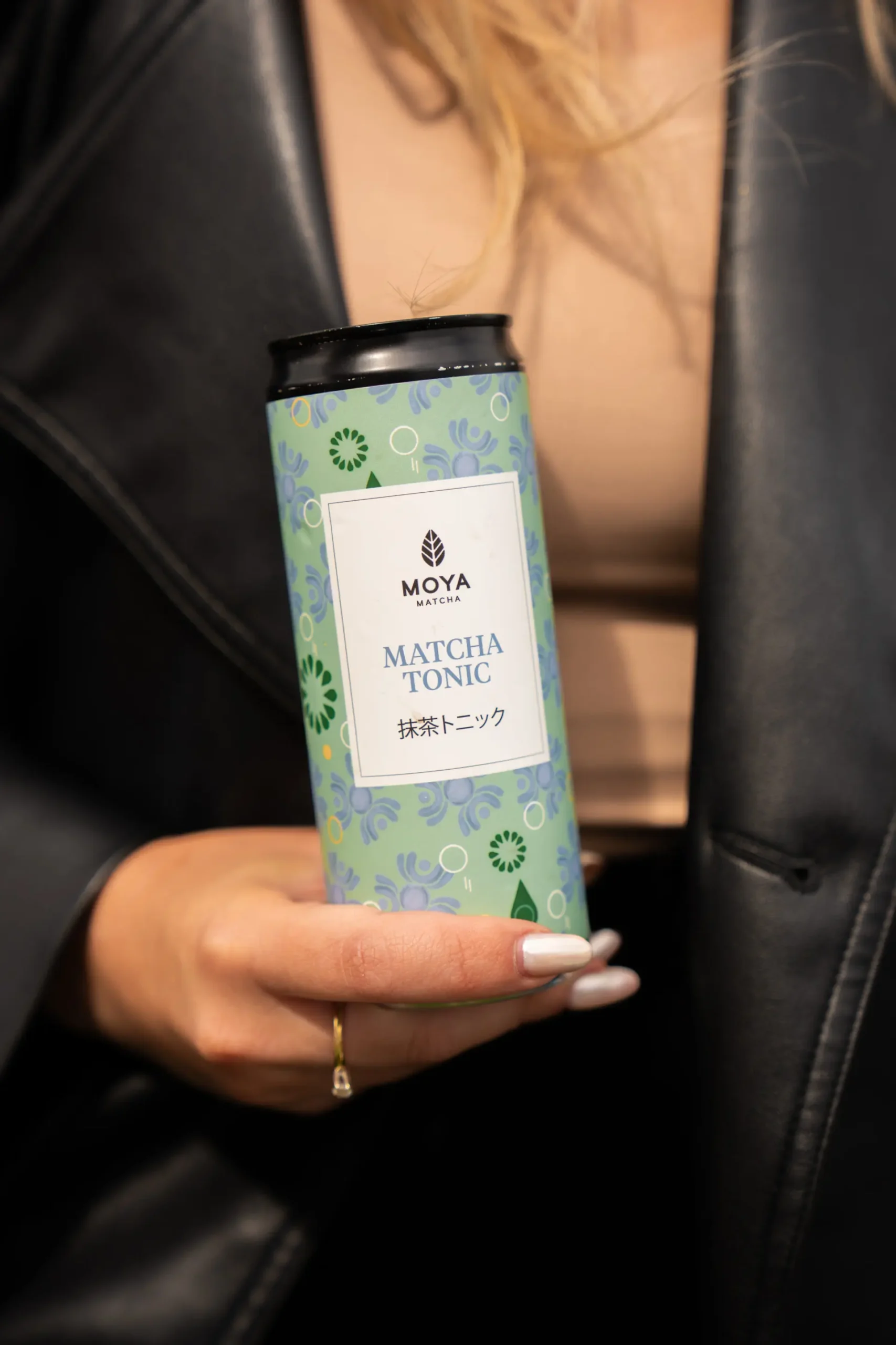 En person i en elegant svart skinnjakke som holder en boks med Moya Matcha Tonic i hånden.
