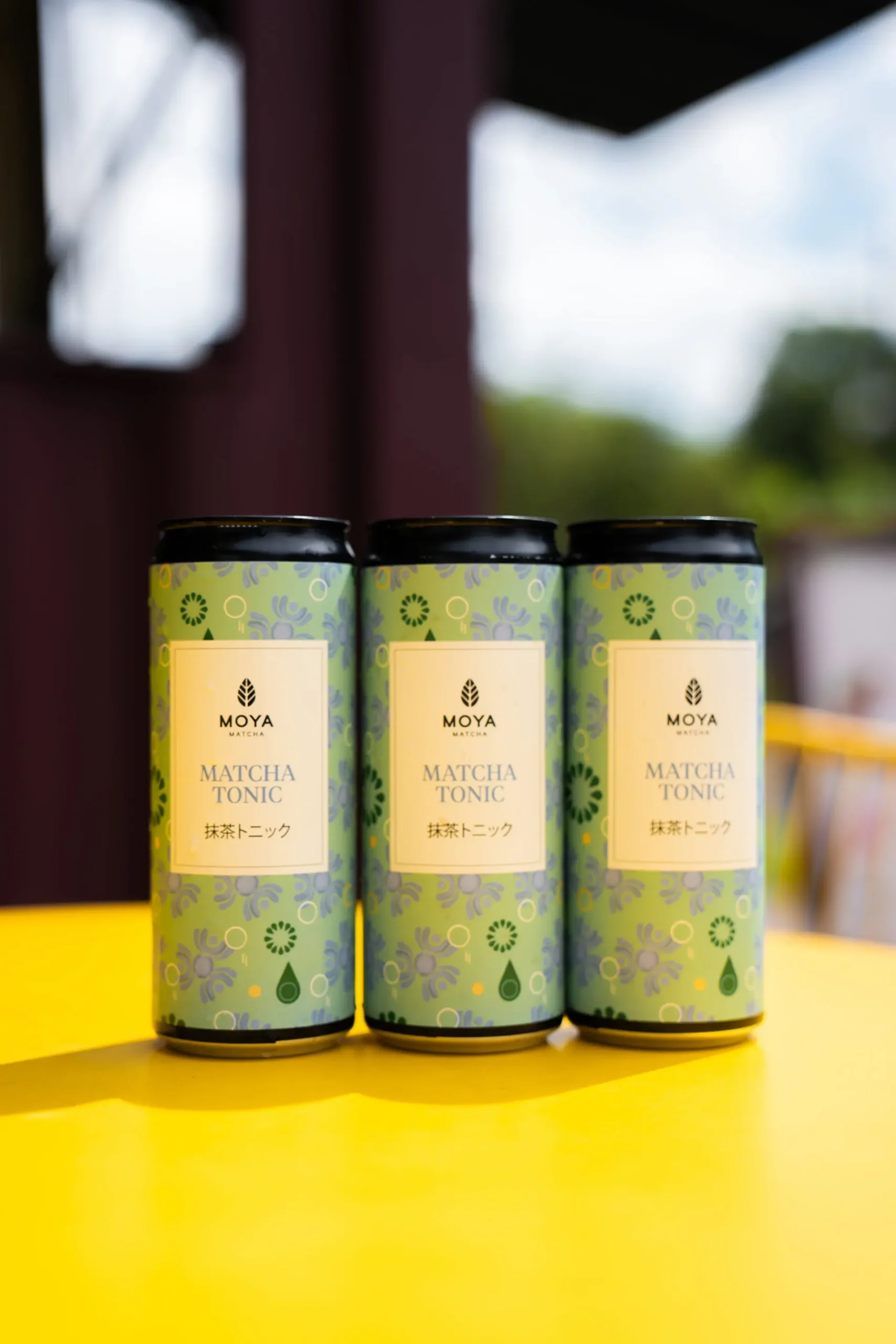 Tre bokser med Moya Matcha Tonic 330ml stående på rad på et gult bord utendørs.